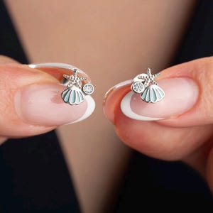 May include: A pair of silver seashell earrings. Each earring features a light blue seashell, a starfish, and a small, clear crystal. The earrings are held up by a person's fingers against a dark background.