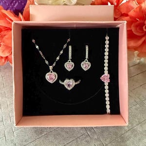 May include: A jewelry set in a pink gift box. The set includes a necklace, earrings, ring, and bracelet, all featuring heart-shaped pink gemstones. The jewelry is silver-toned and set against a black background.
