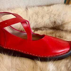 May include: A red leather shoe with a strap and buckle closure. The shoe has a pointed toe and a black sole. The shoe is sitting on a fur surface.