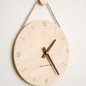 May include: A wooden wall clock with a natural wood finish and black hour markers. The clock face has the text "Enjoy Your Time" printed on it. The clock is hanging from a rope.