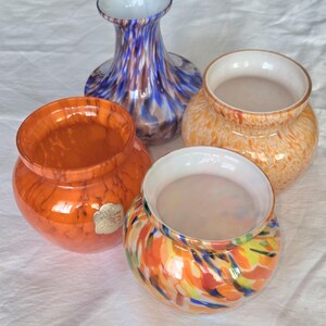 Könnte beinhalten: Eine Sammlung von vier Glasgefäßen, bestehend aus einer hohen blau-weiß gesprenkelten Vase, einer einfarbigen orangefarbenen Vase mit einem kleinen Metallaufkleber, einer hellorangefarbenen gesprenkelten Vase und einer bunten Vase mit orangefarbenen, gelben und blauen Glaswirbeln.