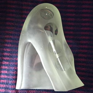 Può includere: Una scultura in vetro satinato traslucido caratterizzata da una forma astratta e curva, con una serie di quattro aperture circolari in vetro trasparente disposte in linea verticale lungo il lato, creando una silhouette liscia e opaca.