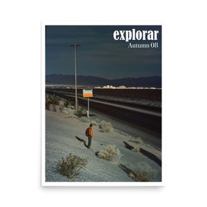 May include: A framed print featuring a desert landscape with a road and a person standing on a hillside. The print has the word "explorar" and "Autumn 08" in white text at the top. The person is wearing an orange jacket and a backpack.
