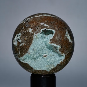 May include: A spherical geode with a rough, brown exterior and a large cavity filled with light blue, crystalline formations. The geode is mounted on a black stand, showcasing its natural beauty and unique texture.