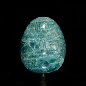 Puede incluir: Una gema pulida en forma de huevo, en tonos turquesa y azul. La piedra tiene una estructura cristalina con fracturas internas visibles, creando una apariencia texturizada. El objeto está sobre un fondo negro.