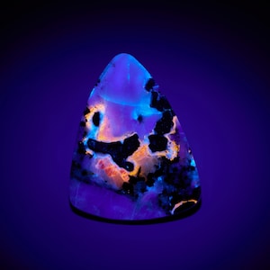 May include: A triangular-shaped gemstone with a vibrant, multi-colored pattern. The stone features a mix of black, orange, and white inclusions against a purple and blue background. The gemstone is illuminated with a blue light.