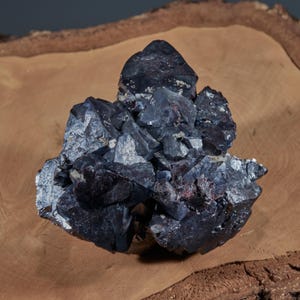 Pode incluir: Um aglomerado mineral escuro e cristalino com uma estrutura complexa e irregular. O mineral exibe uma cor cinza carvão profundo com toques de prata e marrom avermelhado. O espécime está sobre um fundo de madeira natural.