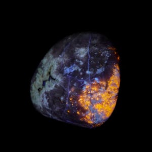 May include: A glowing, rounded rock against a black background. The rock has a dark, mottled surface with patches of blue and green. Bright orange and yellow areas illuminate the right side, with blue veins running through it.