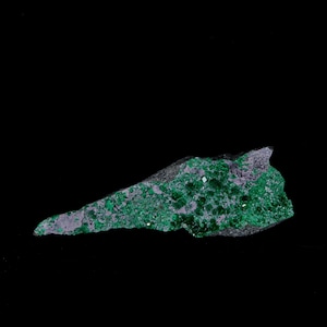 May include: A piece of raw, uncut mineral with a dark gray base and vibrant green crystalline formations. The mineral has a rough, uneven texture and is set against a solid black background.
