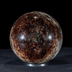 May include: A polished sphere with a complex pattern of dark brown and tan mineral inclusions. The orb sits on a small, silver-colored base, with a black background. The sphere's surface reflects light.