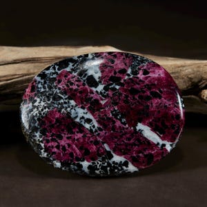 May include: A polished oval gemstone featuring a vibrant mix of deep magenta, stark black, and bright white mineral inclusions. The stone has a smooth, reflective surface with a complex, mottled pattern of contrasting colors throughout.