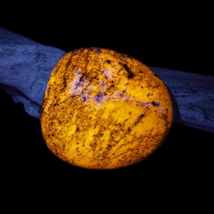 May include: A glowing, rounded rock with a vibrant yellow-orange hue, speckled with dark brown and hints of purple. The rock is set against a black background with a blue-gray wooden element in the background.