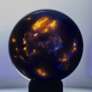 Puede incluir: Una esfera oscura con elementos internos brillantes. La esfera tiene un exterior azul oscuro y negro, con patrones internos de color naranja brillante y amarillo. El objeto está sobre una base negra, y el fondo es un degradado de blanco y azul.