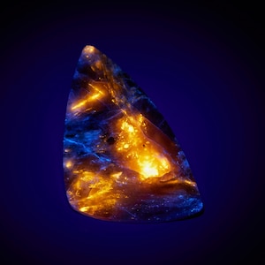 Puede incluir: Una gema pulida de forma irregular, que muestra una vibrante paleta de colores. La piedra presenta tonos naranja fuego y azul brillante, contrastando con un fondo azul oscuro. Los patrones internos se asemejan a vetas brillantes.