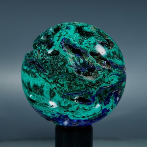 Op de afbeelding: Een bolvormige azuriet- en malachiet-edelsteen. De steen heeft een mix van diepe teal, turquoise en donkerblauwe tinten. Het oppervlak heeft een textuur met natuurlijke formaties en kleine holtes. De edelsteen is op een zwarte standaard gemonteerd.