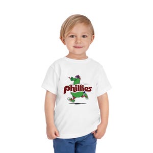 May include: White toddler t-shirt with a green cartoon character running and the word "Phillies" in red letters.
