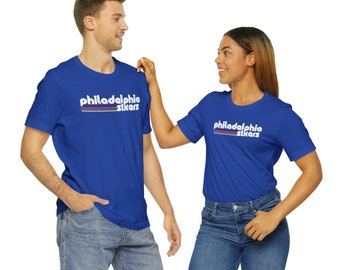 May include: Two people wearing royal blue t-shirts with the text "Philadelphia Sixers" in white with red and blue stripes. The person on the left is wearing blue jeans. The person on the right is wearing blue jeans.