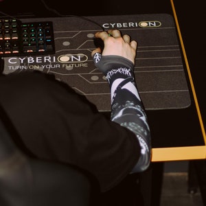Może przedstawiać: Ręka używająca czarnej podkładki pod mysz z tekstem "CYBERION TURN ON YOUR FUTURE" i logo "CYBERION".