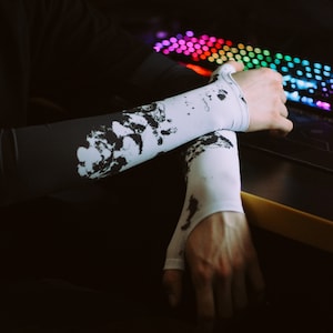 以下が含まれることがあります： 黒のインクの飛沫模様がついた白いアームスリーブを着用した人物。スリーブは両腕に着用され、手首で折り畳まれています。人物は虹色のキーボードが置かれた机に座っています。