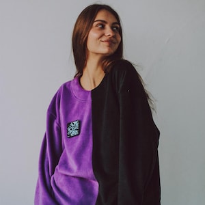 May include: A purple and black sweatshirt with a small graphic design on the left side. The sweatshirt has a crew neck and long sleeves.