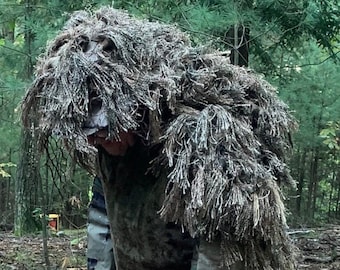 May include: A person wearing camouflage clothing and a ghillie suit, standing in a wooded area with brown leaves and twigs on the ground.