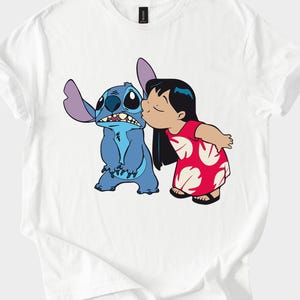 May include: White t-shirt featuring a cartoon illustration of Stitch, a blue alien, being kissed on the cheek by a girl in a red dress. The design is a colorful graphic print, perfect for fans of the animated movie.