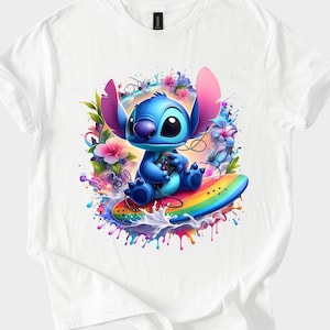 Peut inclure: T-shirt blanc avec un personnage de dessin anim&eacute; bleu tenant une manette de jeu, assis sur une planche de surf arc-en-ciel. Le motif comprend des &eacute;l&eacute;ments floraux et d'&eacute;claboussures color&eacute;s.