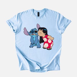 May include: Light blue t-shirt featuring a graphic of Stitch from Lilo & Stitch being kissed by Lilo. Stitch is blue with large ears, and Lilo wears a red dress with white floral patterns. The shirt has rolled-up sleeves.