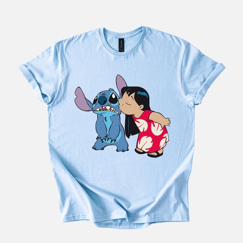 May include: Light blue t-shirt featuring a graphic of Stitch from Lilo & Stitch being kissed by Lilo. Stitch is blue with large ears, and Lilo wears a red dress with white floral patterns. The shirt has rolled-up sleeves.