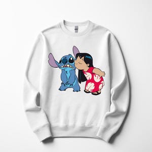 May include: White sweatshirt featuring a graphic of Stitch and Lilo from the Disney movie Lilo & Stitch. Lilo is kissing Stitch on the cheek. Stitch is blue with large ears. Lilo is wearing a red dress with white floral designs.