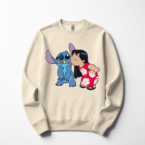 May include: Cream-colored sweatshirt featuring a graphic of Stitch, a blue alien, and Lilo, a girl in a red dress. Lilo is kissing Stitch on the cheek. The design is a cartoon style.