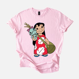 Puede incluir: Camiseta rosa claro con una ilustraci&oacute;n de dibujos animados de una ni&ntilde;a sosteniendo una mu&ntilde;eca y una bolsa verde. La ni&ntilde;a lleva un vestido rojo con estampados de hojas blancas y sandalias azules. La mu&ntilde;eca es azul con un lazo rosa.