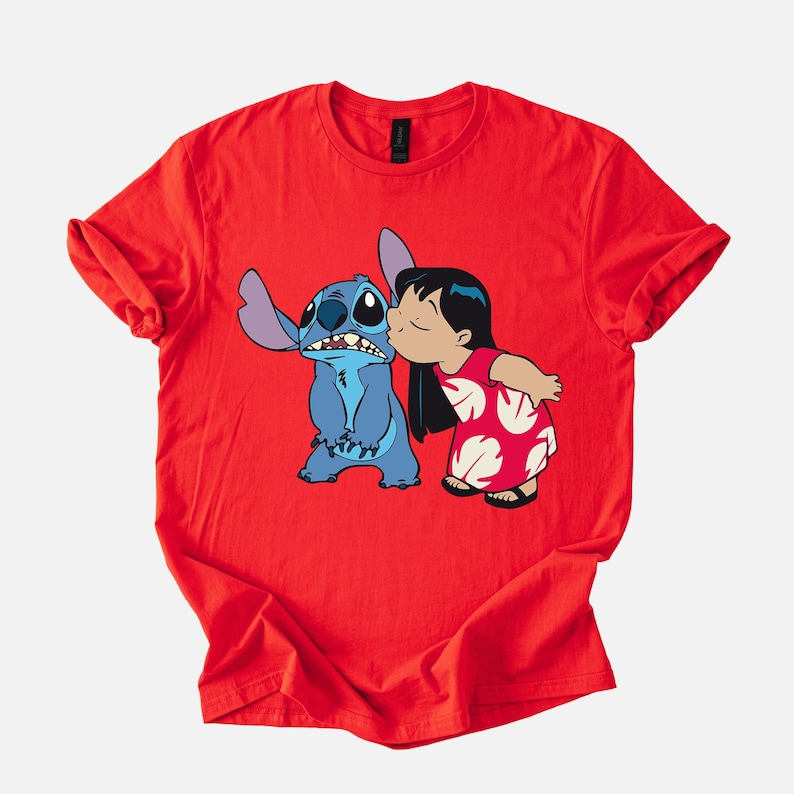 May include: Red t-shirt featuring a graphic of Stitch, a blue alien, being kissed on the cheek by a girl in a red dress. The design is a cartoon style, with bold outlines and bright colors. The shirt is made of a soft material.