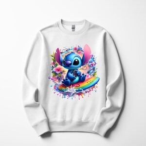 Peut inclure: Sweat-shirt blanc avec un personnage de dessin anim&eacute; bleu sur une planche de surf arc-en-ciel, entour&eacute; de fleurs et d'&eacute;claboussures de peinture. Le personnage a de grandes oreilles. Design ludique et color&eacute;.