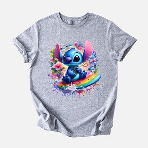 Peut inclure: T-shirt gris clair avec un motif color&eacute; d'un personnage de dessin anim&eacute; bleu aux grandes oreilles, assis sur une planche de surf arc-en-ciel. Le personnage est entour&eacute; de fleurs et d'&eacute;claboussures d'eau.