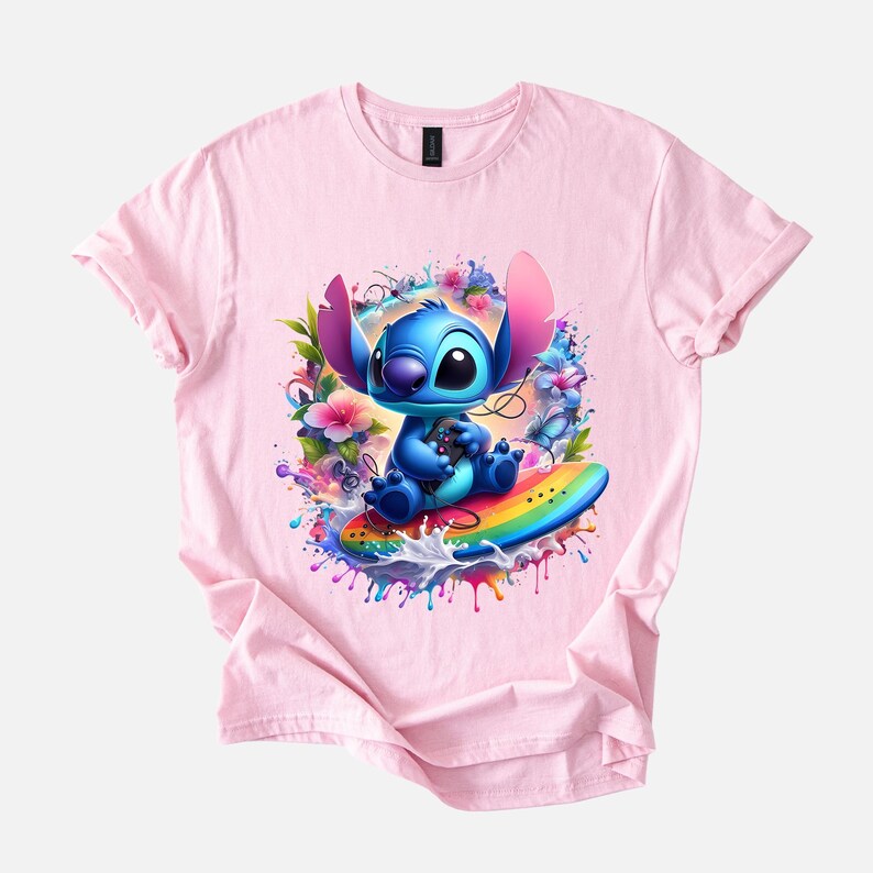 Peut inclure: T-shirt rose clair avec un personnage de dessin anim&eacute; bleu assis sur une planche de surf arc-en-ciel. Le personnage est entour&eacute; de fleurs color&eacute;es et d'&eacute;claboussures d'eau. Le design est vif et d&eacute;taill&eacute;, id&eacute;al pour les fans du personnage.