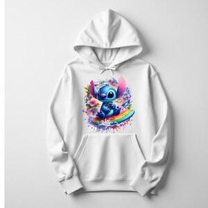 Peut inclure: Sweat &agrave; capuche blanc avec un motif color&eacute; d'un personnage de dessin anim&eacute; bleu sur une planche de surf arc-en-ciel, entour&eacute; de fleurs et d'&eacute;claboussures de peinture. Le sweat &agrave; capuche a une capuche &agrave; cordon et une poche avant.