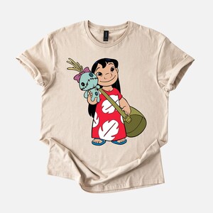 Puede incluir: Camiseta beige con una ilustraci&oacute;n de dibujos animados de una ni&ntilde;a con un vestido rojo con detalles blancos, sosteniendo una mu&ntilde;eca azul y una bolsa verde. La ni&ntilde;a tiene el pelo negro y est&aacute; sonriendo. La camiseta es de material suave.