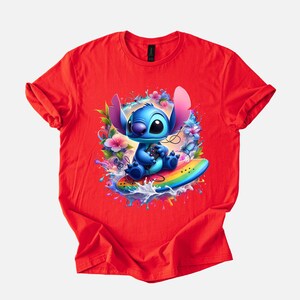 Peut inclure: T-shirt rouge avec un graphisme vibrant d'un personnage de dessin anim&eacute; bleu sur une planche de surf arc-en-ciel, entour&eacute; de fleurs et d'&eacute;claboussures d'eau. Le personnage a de grandes oreilles et tient une manette de jeu.