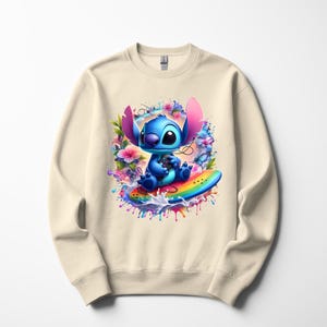 Peut inclure: Sweat-shirt cr&egrave;me avec une illustration vibrante d'un personnage de dessin anim&eacute; bleu sur une planche de surf arc-en-ciel. Le personnage est entour&eacute; de fleurs color&eacute;es et d'&eacute;claboussures de peinture, cr&eacute;ant un design ludique.