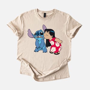 May include: Beige t-shirt featuring a cartoon illustration of Stitch and Lilo from the Disney movie Lilo & Stitch. Lilo is kissing Stitch on the cheek. Stitch is blue with large ears. Lilo is wearing a red dress with white floral patterns.