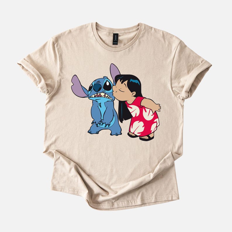 May include: Beige t-shirt featuring a cartoon illustration of Stitch and Lilo from the Disney movie Lilo & Stitch. Lilo is kissing Stitch on the cheek. Stitch is blue with large ears. Lilo is wearing a red dress with white floral patterns.