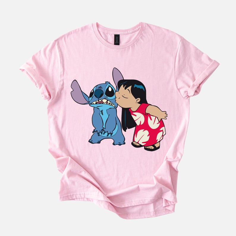 May include: A light pink t-shirt featuring a cartoon graphic of Stitch and Lilo. Stitch is blue, and Lilo is wearing a red dress with white floral designs. Lilo is kissing Stitch on the cheek. The t-shirt has a crew neck and short sleeves.