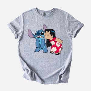 May include: A light gray t-shirt featuring a graphic of Stitch, a blue alien, being kissed by Lilo, a girl in a red dress. The design is a cartoon illustration from the Disney movie Lilo & Stitch.