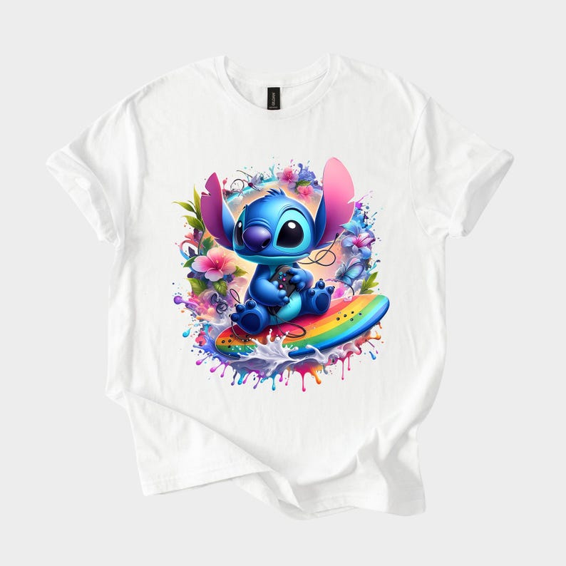 Peut inclure: T-shirt blanc avec un personnage de dessin anim&eacute; bleu tenant une manette de jeu, assis sur une planche de surf arc-en-ciel. Le motif comprend des &eacute;l&eacute;ments floraux et d'&eacute;claboussures color&eacute;s.