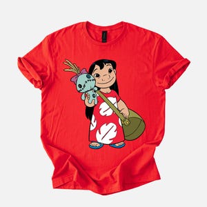 Puede incluir: Camiseta roja con una ilustraci&oacute;n de dibujos animados de una ni&ntilde;a sosteniendo una mu&ntilde;eca y una bolsa verde. La ni&ntilde;a lleva un vestido rojo con estampados blancos en forma de hoja y sandalias azules. La mu&ntilde;eca es azul con un lazo rosa.