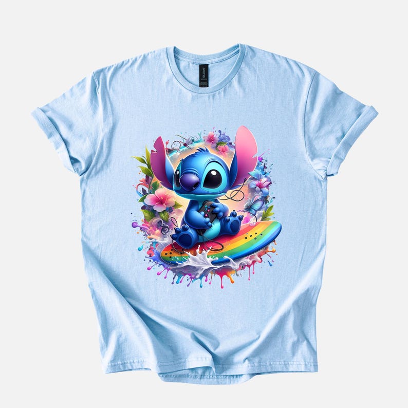 Peut inclure: T-shirt bleu clair avec un motif color&eacute; d'un personnage extraterrestre bleu assis sur une planche de surf arc-en-ciel, entour&eacute; de fleurs et d'&eacute;claboussures de peinture. Le personnage tient une manette de jeu. Le t-shirt a un col rond et des manches courtes.