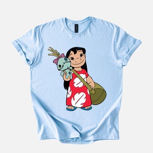 Puede incluir: Camiseta azul claro con una ilustraci&oacute;n de dibujos animados de una ni&ntilde;a con un vestido rojo con dise&ntilde;os blancos en forma de hoja. La ni&ntilde;a sostiene una bolsa verde y una mu&ntilde;eca azul. La camiseta tiene cuello redondo y mangas cortas.