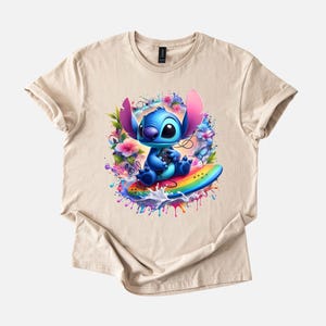 Peut inclure: T-shirt beige avec un motif color&eacute; d'un personnage de dessin anim&eacute; bleu sur une planche de surf arc-en-ciel, entour&eacute; de fleurs et d'&eacute;claboussures de peinture. Le personnage a de grandes oreilles et tient une manette de jeu. Le t-shirt est en mati&egrave;re souple.
