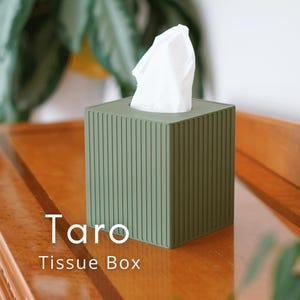May include: A sage green tissue box with a square shape and vertical grooves. White tissues protrude from the top. The text "Taro Tissue Box" is displayed in white. The box is on a wooden surface.